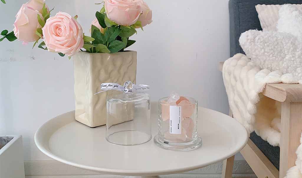 Sunlit cozy corner with pink roses, glass cloche and wax pieces on a round table beside a blanket-draped sofa in a minimal living room.